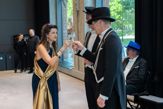 A woman in a dark dress with a gold sash and three men in suits, two with top hats, toast with champagne flutes.