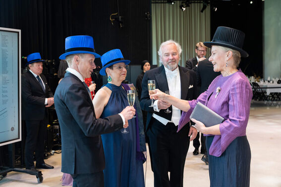 Four people toast with champagne at a formal event. Two wear bright blue hats, one wears a black hat, and one is in formal attire.
