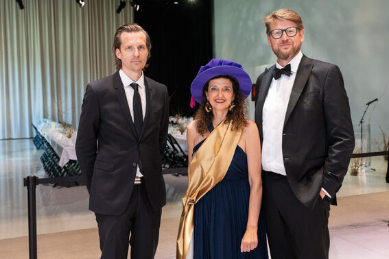 Three people dressed formally with one wearing a unique blue hat and gold sash. A decorated banquet table is in the background.