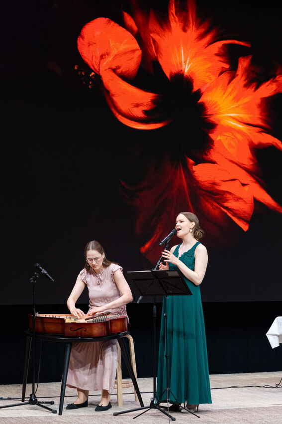 Two women performing: one plays a wooden string instrument while another sings under a backdrop of red abstract art.