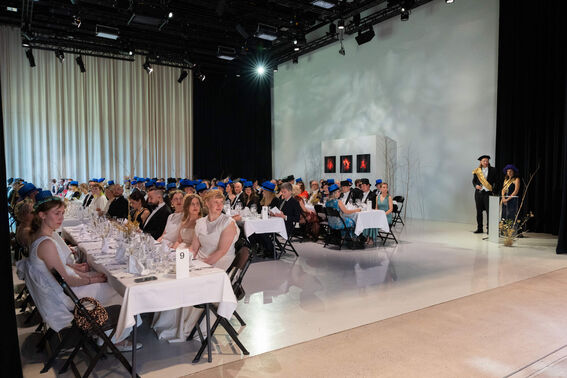 A graduation event with people seated at tables, wearing blue caps. Two presenters stand on the right side of the room.