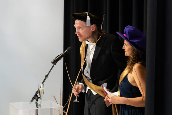 Two people in formal attire with hats, one holding a champagne flute near a microphone.