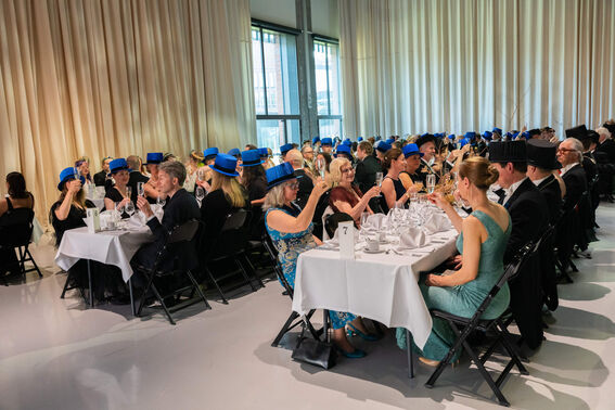 A formal gathering with people wearing blue hats, dressed elegantly. They are sitting at tables and raising glasses for a toast.