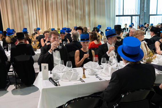 A formal gathering with people seated at tables, wearing black and blue ceremonial hats.
