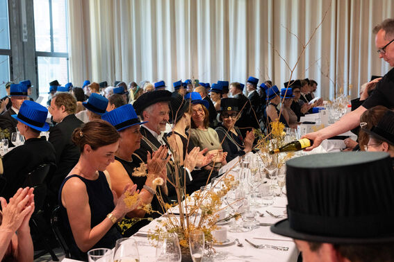 Formal event attendees with blue hats clapping, seated at a long white table with glasses and yellow flower decorations