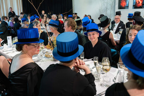 A formal event with people wearing black attire and blue academic hats sitting at a white-clothed table.