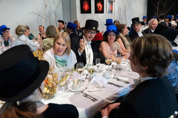 A formal gathering with people in suits and dresses, wearing black and blue hats, seated around a table with drinks.