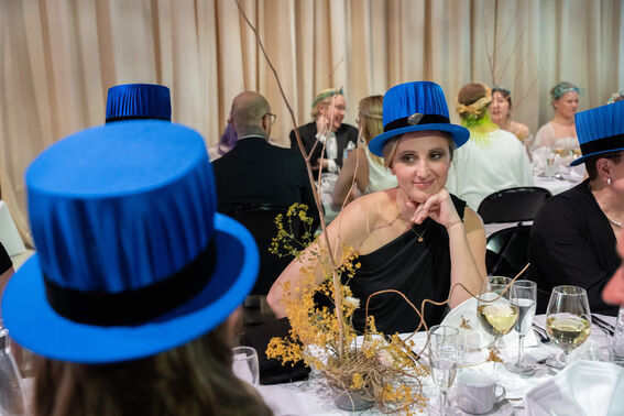 People seated at a banquet wearing blue hats with black bands, elegant table setting with flowers and drinks.