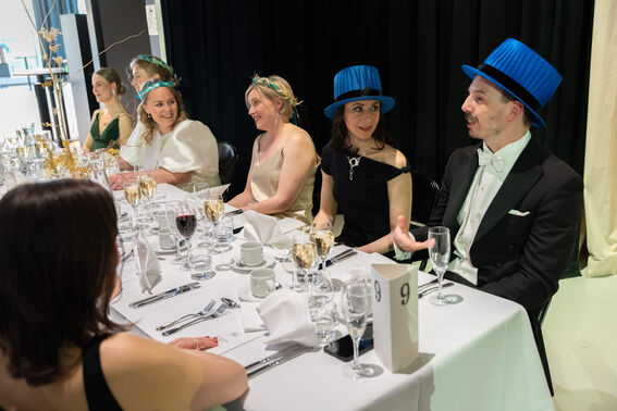 People seated at a formal dinner table. Two guests wear blue top hats. Table number 9 is visible.