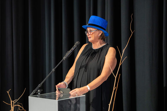 A woman wearing a vibrant blue academic top hat, standing at a clear podium and speaking into a microphone.