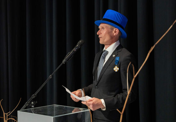 A person in formal attire with blue insignia and hat speaking at a podium, with black curtains in the background.