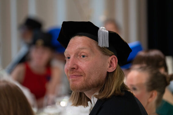 A person wearing a black academic cap with white tassel, attending an event with blurred attendees in the background.