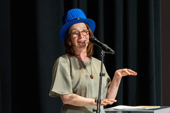 A person in a blue hat speaks at a microphone, wearing a pendant necklace and light gray clothing.