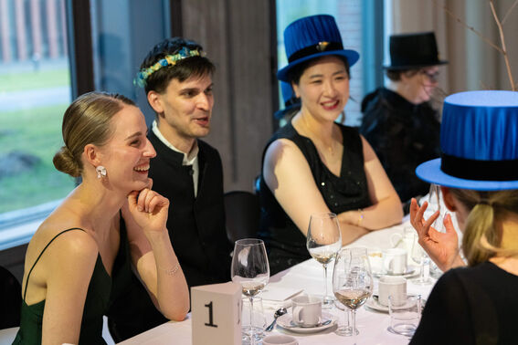Group of people at a formal event, seated at a table with wine glasses. Some are wearing blue hats and black outfits.