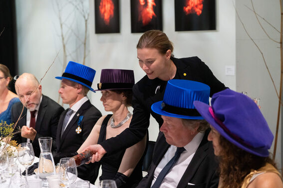 People wearing formal attire and colourful hats sit at a decorated dining table. Art pieces are visible on the wall.