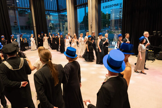 A group of people in formal attire dancing in a hall with large windows, some wearing blue top hats.