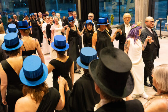 A group of people in formal attire participating in a dance. Many are wearing blue top hats.