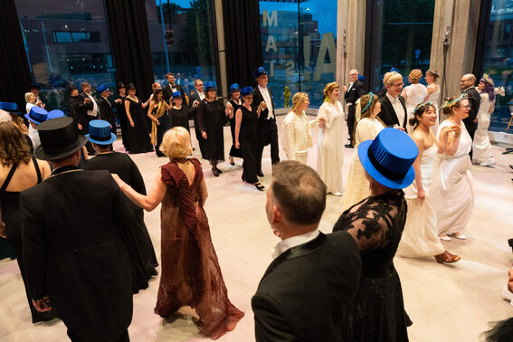 Formal dance event indoors with participants in evening wear; some wear blue hats. Large windows in background.