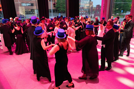 People in formal attire and blue hats dancing at an indoor event, with large windows showing a view of buildings outside.