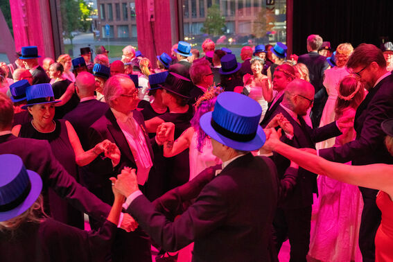 A group of people in formal attire dancing, many wearing blue top hats. The room is illuminated with pink lighting.