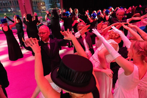 A group of people in formal attire and white gloves dancing indoors, with several wearing blue hats.