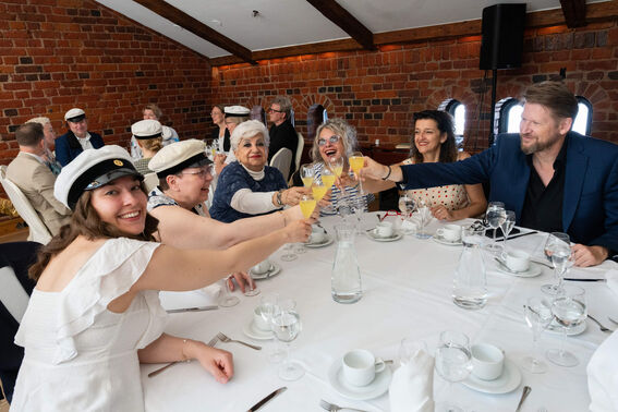 People seated around a table with a white cloth, making a toast with drinks. Brick wall background, wooden beams on the ceiling.