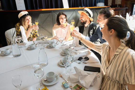 Five people sitting at a restaurant table toasting with drinks. Table set with glasses, coffee cups, and white napkins.