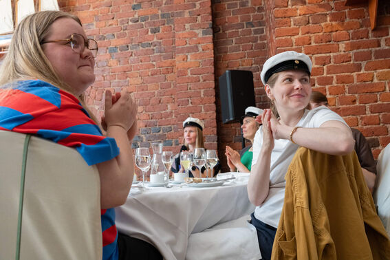 People at a table in a brick-walled room, wearing white caps, clapping. Table has wine glasses and plates.