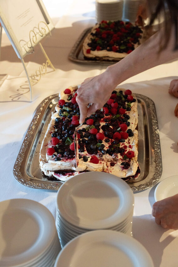 A hand cutting a pavlova topped with mixed berries at a table, next to a stack of white plates.