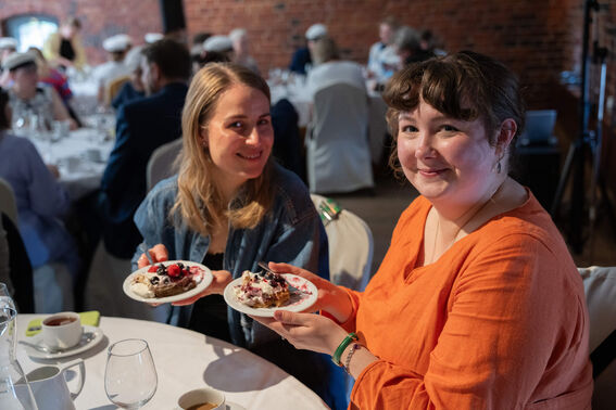 Two people holding plates of dessert at a formal gathering, seated at a table with a white tablecloth.