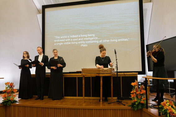 Four musicians performing on stage with a projected quotation by Plato. Floral decorations are on both sides.