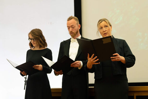 Three people dressed formally holding black folders while standing in front of a white and green screen.