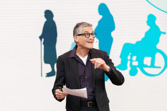 A man in a black suit speaks while holding papers. Silhouettes of various figures are depicted in the background.