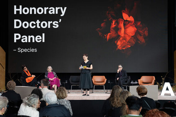 A panel discussion with four participants on stage, chairs, a large screen displaying 'Honorary Doctors' Panel - Species'.