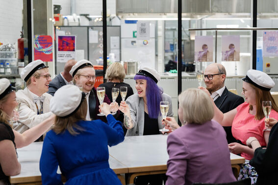 People in festive hats raise glasses in a toast around a table inside a modern room, some with colourful clothing.