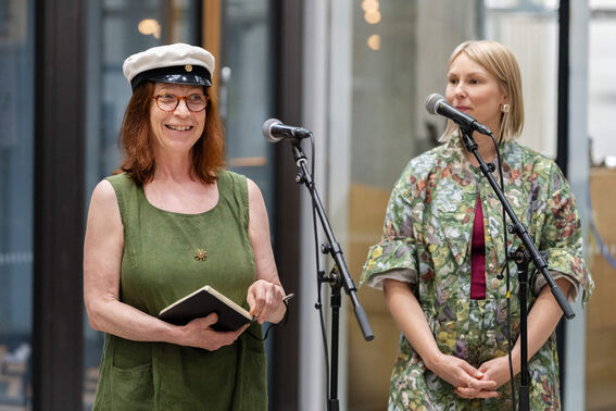Two women standing and speaking into microphones. The left woman holds a book and wears a white hat; the right wears a floral dress.