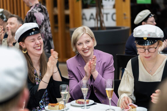 Group of people in formal attire clapping. Lady in purple jacket, white caps. Glasses of champagne and plates on table.