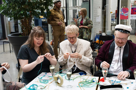 People sitting around a table, working with ribbons and various crafts, with glasses of champagne on the table.