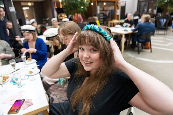 A woman with long hair wearing a blue and green headband sits at a table in a crowded indoor event.
