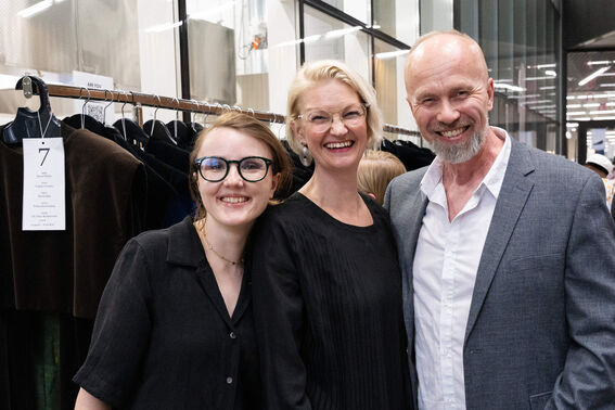 Three people posing in front of a clothing rack indoors. One man in a grey jacket, two women in black attire.