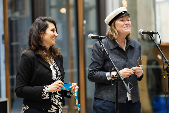 Two women speaking into microphones, one holding a wreath and the other wearing a student cap.