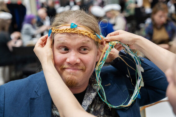 Person is getting a woven floral crown placed on their head at a celebration. Others are visible in the background.