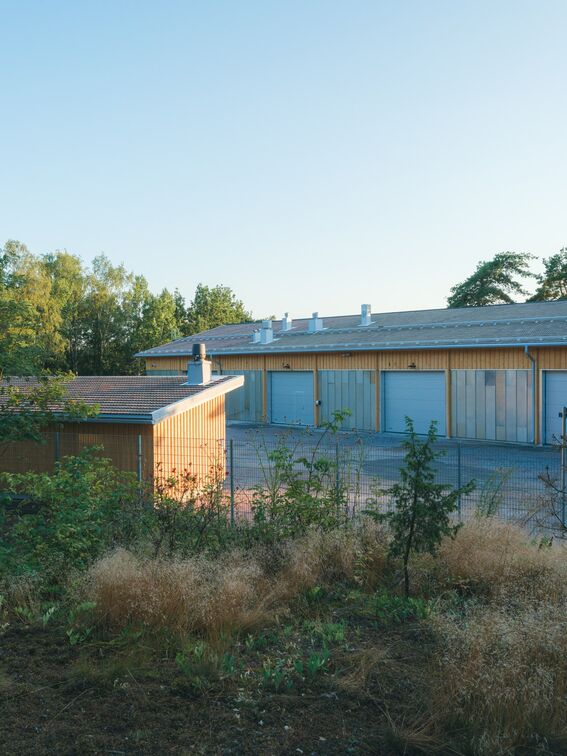 Wooden, storage buildings with grey roofs behind a fence and vegetation in the foreground.