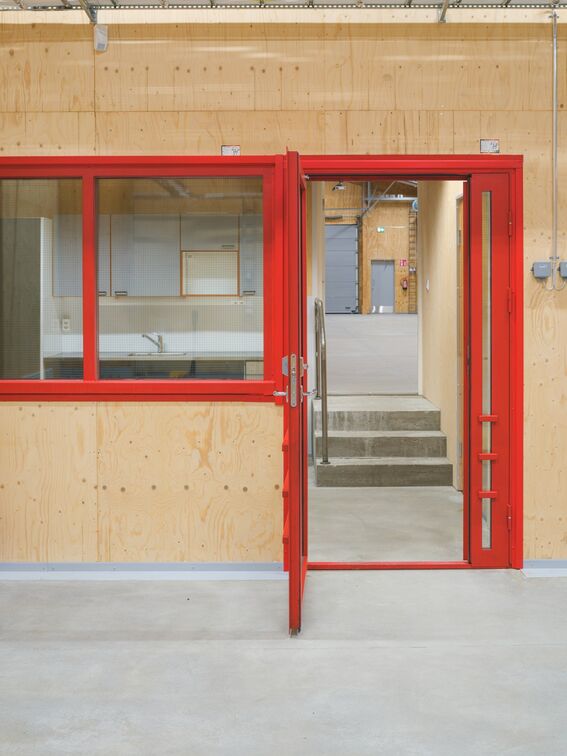 A red-framed glass door and window in a light wooden wall. The door is open, showing stairs leading to an industrial area.