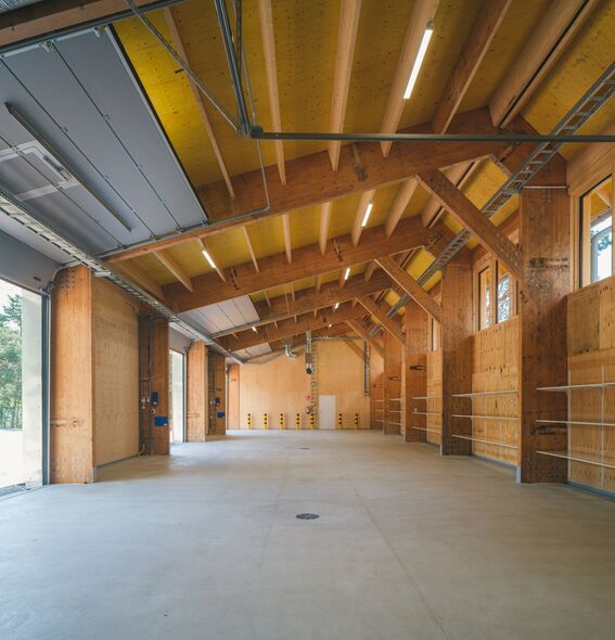 A spacious warehouse with wooden beams, large open doors, and concrete floor. The ceiling is lined with bright lights.