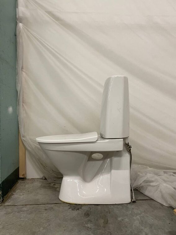 A white toilet in a room with a green wall on the left and a white sheet in the background, placed on a concrete floor.