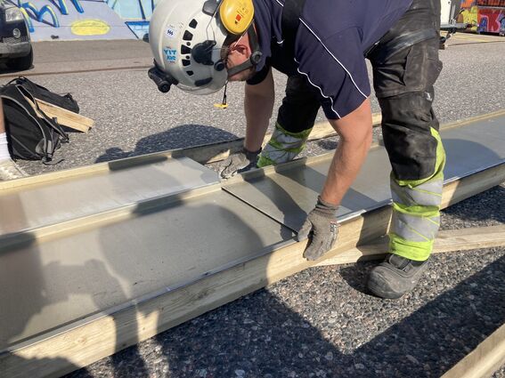 A worker in protective gear is looking at aluminum panels on wooden frames outdoors. Graffiti and vehicles are visible in the background.