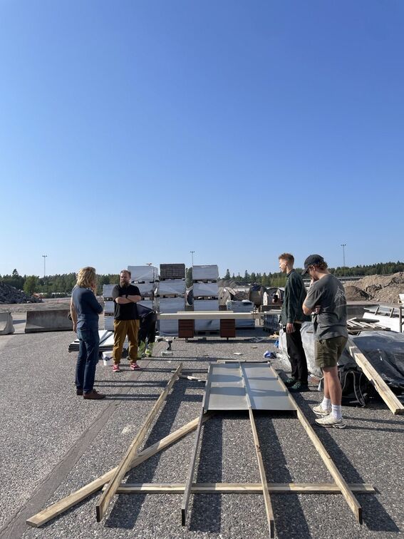 Several people standing beside construction materials on a sunny day in an industrial area with a clear blue sky.