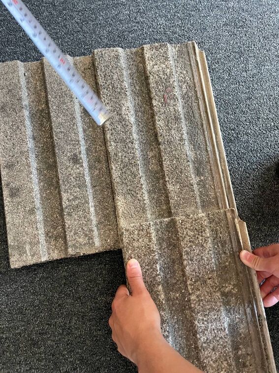 Two hands holding roof tiles. A measuring tape is placed next to them on a dark textured surface.