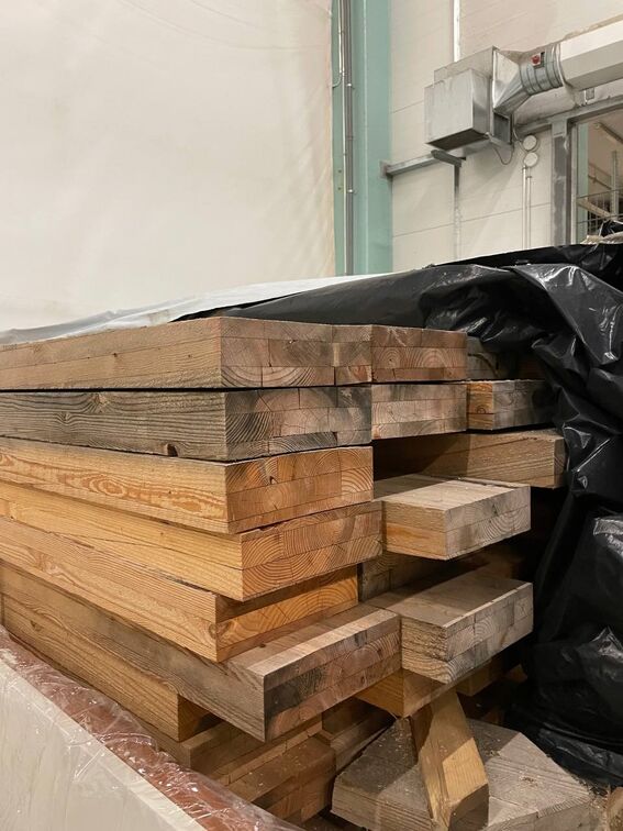 Large stack of glulam wrapped in plastic on one side, with industrial pipes visible in the background.